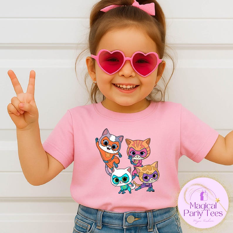 May include: A pink t-shirt featuring a cartoon graphic of four cat characters with superhero masks. The child in the image is wearing heart-shaped sunglasses and blue jeans. The t-shirt has the text "Magical Party Tees" on the bottom right.