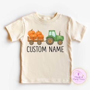 May include: Cream-coloured t-shirt with a green tractor pulling a wooden cart filled with orange pumpkins. The text "CUSTOM NAME" is printed below the tractor. A seasonal design for children's clothing.