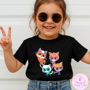 May include: A black t-shirt featuring a colorful graphic of four cartoon cats wearing superhero masks and costumes. The cats are in various poses, with vibrant colors like orange, pink, and blue. The shirt is worn by a child.