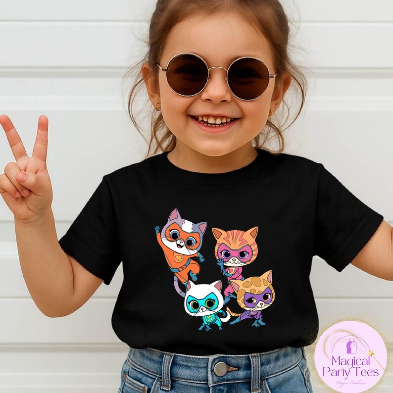May include: A black t-shirt featuring a colorful graphic of four cartoon cats wearing superhero masks and costumes. The cats are in various poses, with vibrant colors like orange, pink, and blue. The shirt is worn by a child.
