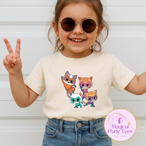 May include: A cream-colored t-shirt featuring a colorful graphic of four cartoon cat characters. The cats are in various poses, with orange, blue, and purple accents. The shirt has the text "Magical Party Tees" on a pink circle.