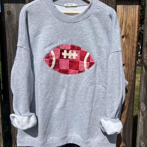 May include: A light gray crewneck sweatshirt featuring a large, central applique of a football made from red and white checkered fabric patches. The text "game day" is embroidered in white cursive script above the football graphic.