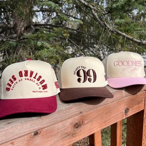 Op de afbeelding: Drie beige baseballpetten met verschillende geborduurde ontwerpen. De eerste pet heeft een donkerrode klep en zegt "Faith as small as a mustard seed" in bruine tekst. De tweede pet heeft een bruine klep en zegt "He left the 99" in bruine tekst. De derde pet heeft een roze klep en zegt "Goodness" in bruine tekst.