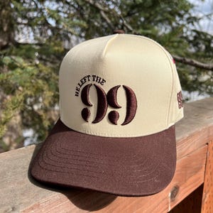 Op de afbeelding: Een beige en bruine baseballpet met de tekst "He Left The 99" geborduurd op de voorkant.