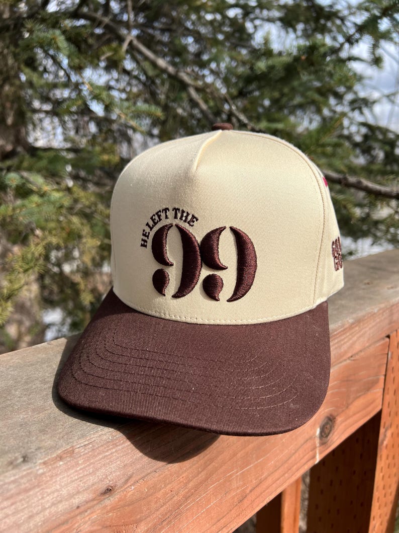 Op de afbeelding: Een beige en bruine baseballpet met de tekst "He Left The 99" geborduurd op de voorkant.