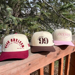 Op de afbeelding: Drie baseballpetten met verschillende ontwerpen. De eerste pet is rood en wit met de tekst "Mustard Seed Faith as small as a" en "Matthew 17:20". De tweede pet is bruin en wit met de tekst "Heaft the 99". De derde pet is roze en wit met de tekst "Live for the Goodness".