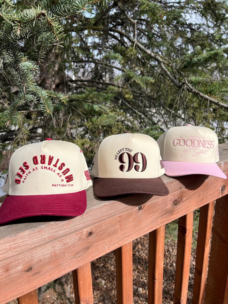 Op de afbeelding: Drie baseballpetten met verschillende ontwerpen. De eerste pet is rood en wit met de tekst "Mustard Seed Faith as small as a" en "Matthew 17:20". De tweede pet is bruin en wit met de tekst "Heaft the 99". De derde pet is roze en wit met de tekst "Live for the Goodness".