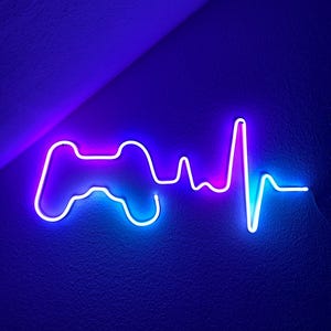 Puede incluir: Letrero de neón con forma de mando de videojuegos y latido del corazón. El letrero brilla con un degradado de colores, pasando del rosa al azul. El letrero está montado en una pared azul oscuro.