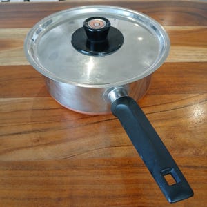 May include: A stainless steel saucepan with a clear lid and a black handle. The lid has a black knob. The saucepan is sitting on a wooden surface. Kitchenware.