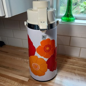 May include: Vintage white and floral patterned beverage dispenser with a cream-colored top and spout. The dispenser features a silver band and handles, with a "Whale" logo. The floral design includes orange and red flowers.