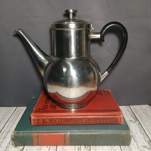 May include: A shiny, silver-colored coffee pot with a black handle and spout, resting on top of two antique books with red and green covers. The coffee pot has a rounded body and a lid.