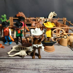 May include: A collection of toy cowboy figures, horses, and a wagon. The figures wear cowboy hats, vests, and hold guns. The scene includes a wooden fence, an anvil, and a skeleton of an animal, creating a Wild West theme.