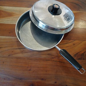 May include: A stainless steel frying pan with a lid, set on a wooden surface. The pan has a black handle and the lid has a black knob. The pan is open, and the lid is slightly ajar.