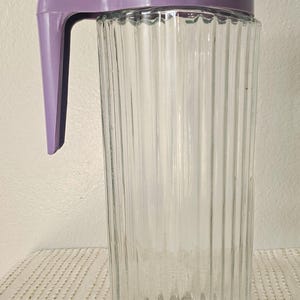 Puede incluir: Una jarra de vidrio transparente con detalles acanalados verticales. Cuenta con un asa y tapa de plástico morado. La jarra tiene forma rectangular con base triangular, adecuada para servir bebidas.