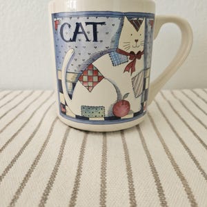 May include: White ceramic mug with a cat illustration. The mug features a seated cat with patchwork details, a red bow, and an apple. The word "CAT" is printed at the top. The mug has a curved handle and is set on a striped surface.