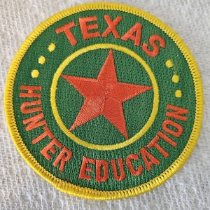 Może przedstawiać: Naszywka haftowana w kolorze zielonym i żółtym z czerwoną gwiazdą w środku. Wokół gwiazdy napis "TEXAS HUNTER EDUCATION".
