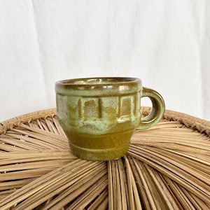 May include: A handmade ceramic mug with a small handle. The mug is a muted olive green colour with a textured, ribbed design around the body. The mug sits on a woven, natural-coloured placemat.