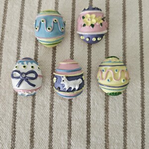 May include: Five decorative Easter egg ornaments in pastel colors. Each egg features unique designs, including floral patterns, bows, and wavy lines. The eggs are painted in shades of blue, pink, yellow, and white, with intricate details.