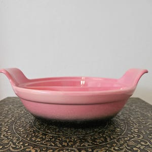 Puede incluir: Un cuenco de cerámica rosa con dos asas curvas. El cuenco tiene un acabado liso y brillante y una forma rectangular poco profunda. El cuenco está sobre una mesa decorativa.
