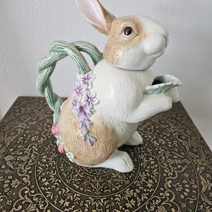 May include: A ceramic bunny teapot with a brown and white body, holding a leaf-shaped spout. The teapot features a green vine handle and is adorned with purple flowers. The teapot is sitting on a decorative, dark brown surface.