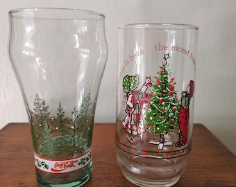 Puede incluir: Dos vasos de Navidad de Coca-Cola vintage. Un vaso presenta una escena de invierno con árboles verdes y un logotipo de Coca-Cola rojo. El otro vaso presenta un árbol de Navidad con dos niños decorándolo. El texto en el vaso dice "Christmas is here... the nicest time".