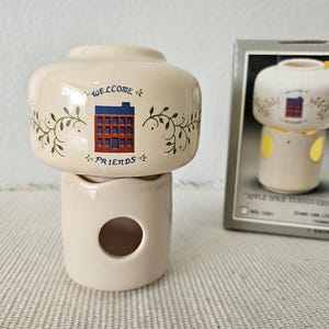 May include: Cream-colored ceramic oil warmer with a house design and the words "Welcome Friends." The warmer has a cylindrical base with a circular opening and a top with a rounded shape. A box with a similar warmer is in the background.