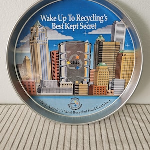 May include: A round metal tray with a cityscape design. The tray features a blue sky background with the text "Wake Up To Recycling's Best Kept Secret." A steel drum is in the center. The tray is labeled "Steel: America's Most Recycled Food Container."