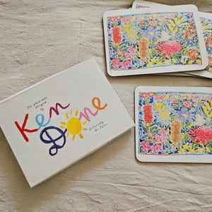 Może przedstawiać: Zestaw sześciu podkładek zaprojektowanych przez Kena Done'a, z kolorowym wzorem polnych kwiatów. Podkładki są prostokątne z białym obramowaniem i napisem "WILDFLOWERS". Pudełko jest białe z nazwiskiem projektanta i ilustracją słońca.