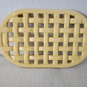 May include: Oval-shaped, pale yellow ceramic trivet with a woven basket design. The trivet is made of ceramic and is designed to protect surfaces from hot dishes. The trivet measures approximately 10 inches long and 7 inches wide.