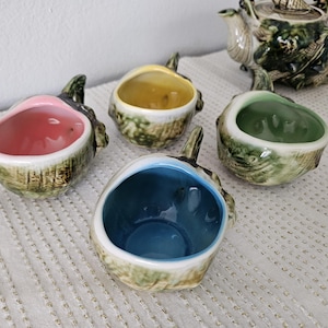 May include: Four ceramic bowls with a unique eggplant design, each with a different interior color: pink, yellow, green, and blue. The bowls have a textured, green and brown exterior. A matching teapot is in the background.
