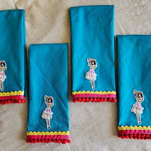 以下が含まれることがあります： カクテルグラスを持った女性が刺繍された、ターコイズブルーのキッチンクロス4枚セット。各クロスは、黄色と赤の縁取りに赤いポンポンが付いています。
