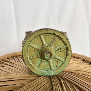 Puede incluir: Una pieza decorativa de cerámica con forma de rueda de carro. La rueda es de color verde apagado con una superficie texturizada y un cubo central. La pieza tiene un aspecto rústico y envejecido, lo que sugiere una estética vintage o antigua. La rueda está sobre una superficie tejida.