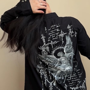 Peut inclure: Chemise noire à boutons avec un motif d'ange détaillé dans le dos. L'ange est représenté avec des ailes et une épée, entouré d'écritures et d'éléments décoratifs. La chemise a des manches longues.