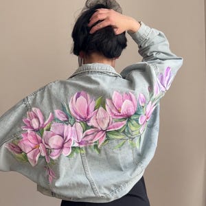 May include: Light blue denim jacket featuring a hand-painted floral design. The back of the jacket is adorned with large pink magnolia blossoms and green leaves. The jacket has a classic collar, button closures, and long sleeves.