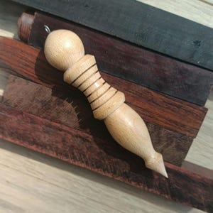 Puede incluir: Un péndulo de madera clara con una parte superior redondeada y una parte inferior puntiaguda. El cuerpo del péndulo tiene varias líneas horizontales talladas. El fondo presenta varias tablas de madera en tonos marrones y negros.