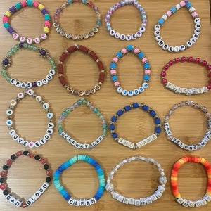 May include: A collection of colorful beaded bracelets with various words spelled out in white letter beads. The bracelets feature a variety of colors, including pink, blue, yellow, and red. Some bracelets have gold accents.
