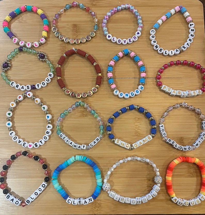 May include: A collection of colorful beaded bracelets with various words spelled out in white letter beads. The bracelets feature a variety of colors, including pink, blue, yellow, and red. Some bracelets have gold accents.