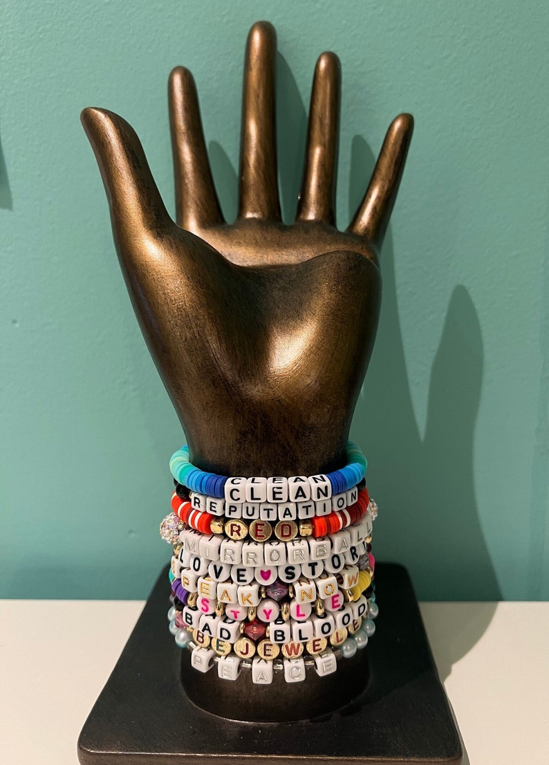 May include: A bronze-colored hand sculpture holds a stack of beaded bracelets. The bracelets feature white letter beads spelling out words like "CLEAN," "REPUTATION," and "PEACE." The bracelets are in various colors, including red, blue, and gold.