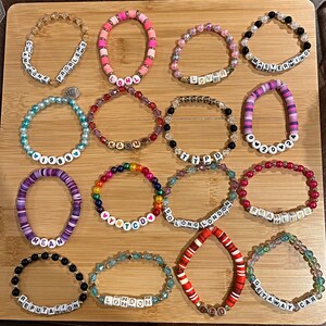 May include: A collection of beaded bracelets in various colors and designs. Some bracelets feature letter beads spelling out words like "Champagne," "Lover," and "Fearless." The bracelets are arranged on a wooden surface.