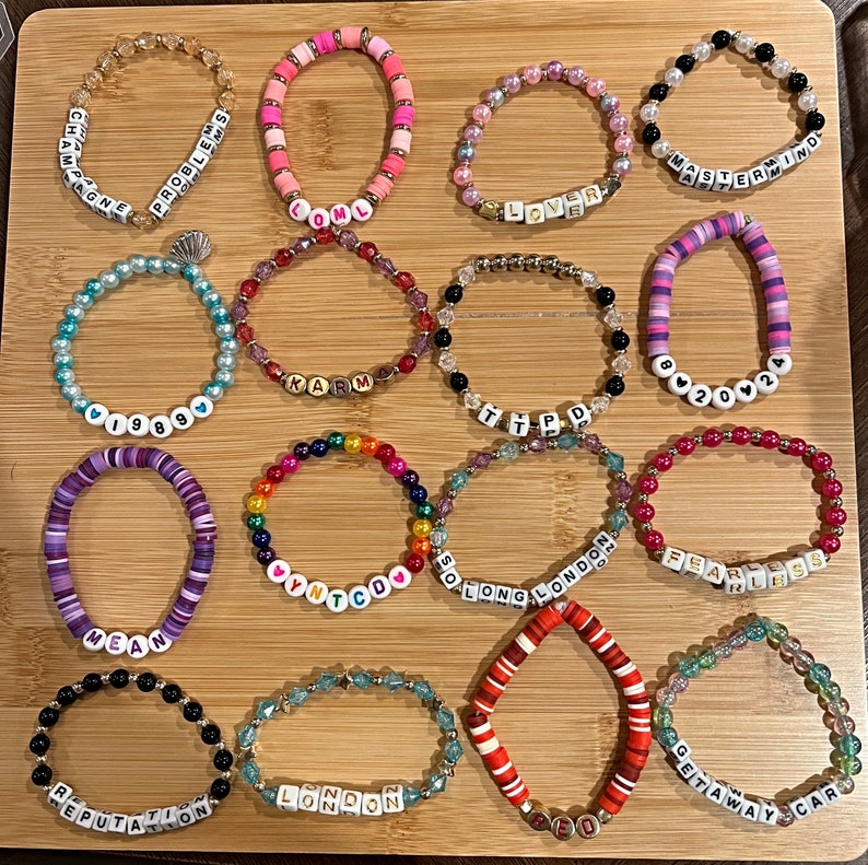 May include: A collection of beaded bracelets in various colors and designs. Some bracelets feature letter beads spelling out words like "Champagne," "Lover," and "Fearless." The bracelets are arranged on a wooden surface.