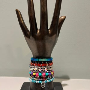 May include: A bronze hand sculpture displays a collection of colorful beaded bracelets. The bracelets feature various colors, including blue, red, white, and purple, with heart and peace sign charms. The bracelets are stacked on the hand.