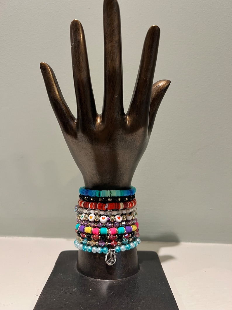 May include: A bronze hand sculpture displays a collection of colorful beaded bracelets. The bracelets feature various colors, including blue, red, white, and purple, with heart and peace sign charms. The bracelets are stacked on the hand.