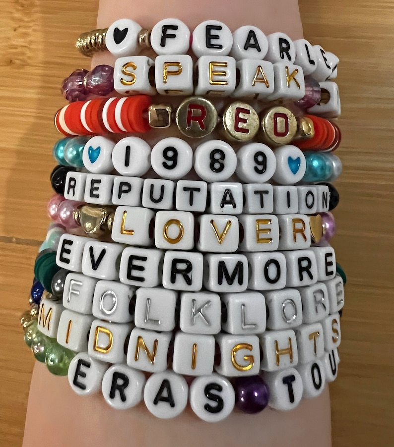 Puede incluir: Una pila de pulseras de cuentas blancas con letras doradas que deletrean "FEARLESS", "SPEAK", "RED", "1989", "REPUTATION", "LOVER", "EVERMORE", "FOLKLORE", "MIDNIGHTS" y "ERAS TOUR". Las pulseras presentan cuentas de acento en rojo, rosa y dorado.
