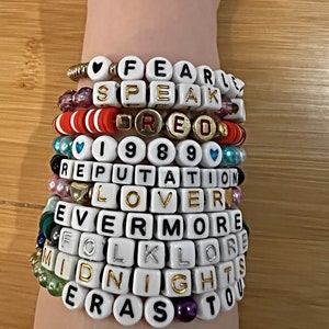 May include: A stack of beaded bracelets spelling out words like "FEARLESS," "SPEAK," "RED," "1989," "REPUTATION," "LOVER," "EVERMORE," "FOLKLORE," "MIDNIGHTS," and "ERAS TOUR." The bracelets are white with black or gold lettering.