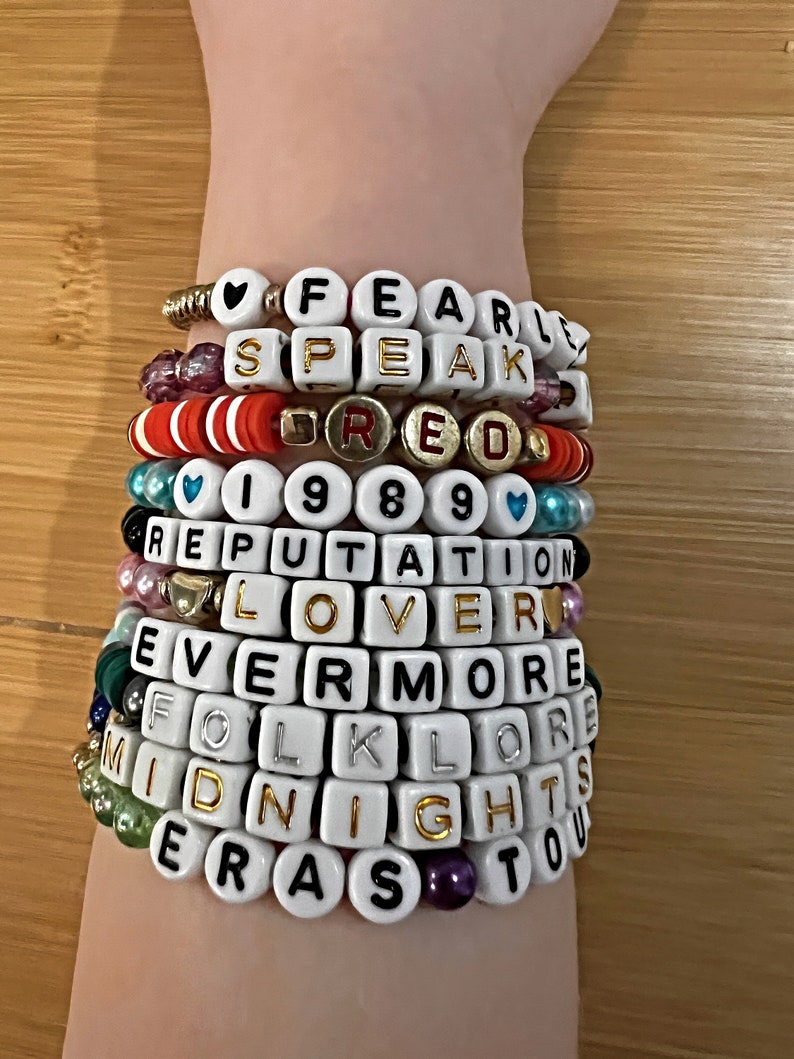 May include: A stack of beaded bracelets spelling out words like "FEARLESS," "SPEAK," "RED," "1989," "REPUTATION," "LOVER," "EVERMORE," "FOLKLORE," "MIDNIGHTS," and "ERAS TOUR." The bracelets are white with black or gold lettering.