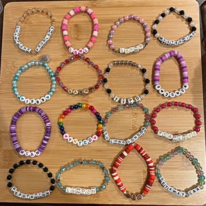 Puede incluir: Una colecci&oacute;n de pulseras de cuentas en varios colores y estilos, con cuentas de letras que deletrean palabras como "Champagne", "Lover" y "Fearless". Las pulseras est&aacute;n dispuestas sobre una superficie de madera.