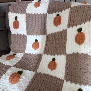 May include: A chunky knit blanket featuring a checkered pattern of white and taupe squares. Each white square contains a textured orange pumpkin motif with a small green stem. The blanket is constructed from thick, soft yarn in a repeating grid.