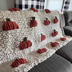 May include: A cream-colored, chunky knit blanket adorned with multiple red pumpkin appliqués with green stems. The blanket is draped over a gray sofa, creating a cozy, autumn-themed aesthetic. The pumpkins are evenly spaced across the blanket.