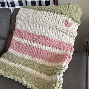 May include: A chunky knit blanket in shades of white, pink, and green. The blanket features a heart-shaped accent and is draped over a gray sofa. The cozy, handmade blanket is perfect for snuggling up.