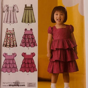May include: Simplicity sewing pattern 2709 for girls' dresses, sizes A 1/2, 1, 2, 3, and 4. The pattern features various dress designs, including tiered and smocked styles. The image shows a young girl wearing a tiered dress. The pattern's brand name is Simplicity.
